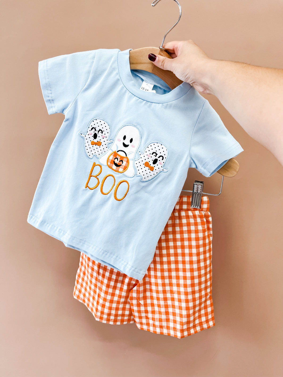 A hand holds the Boo Buddies Halloween Set by Tiny Tot Bundles: a light blue T-shirt with three embroidered ghosts and BOO text, paired with orange and white checkered shorts, against a tan background.