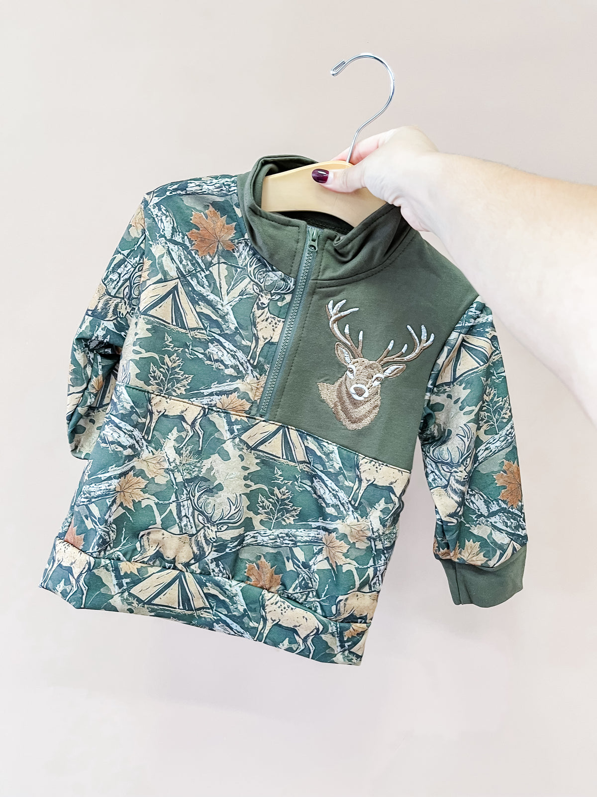 A hand displays the Little Hunter Buck Pullover by Tiny Tot Bundles, a green and tan toddler sweatshirt with a woodland pattern, zipper front, and an embroidered deer head patch on the chest.