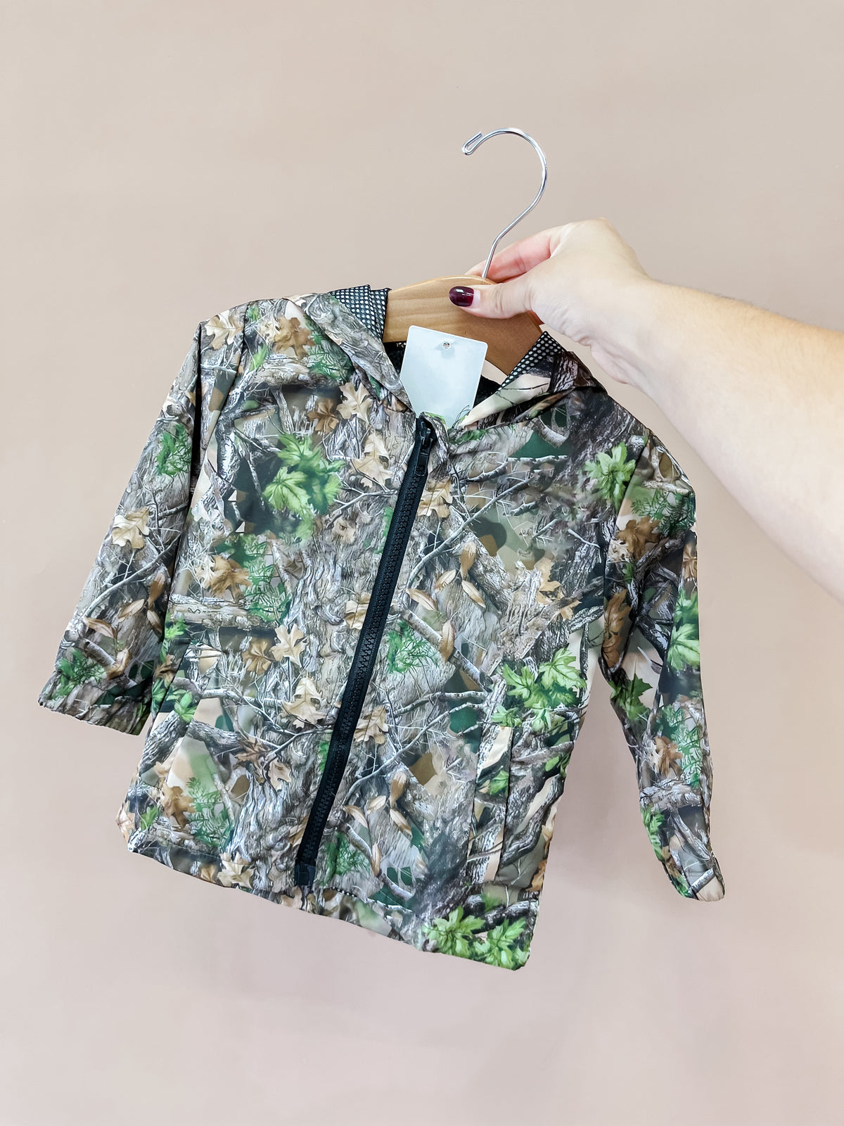 A hand holds the Tiny Tot Bundles Forest Trail Zip Jacket—featuring a camouflage pattern—on a wooden hanger against a plain beige background.