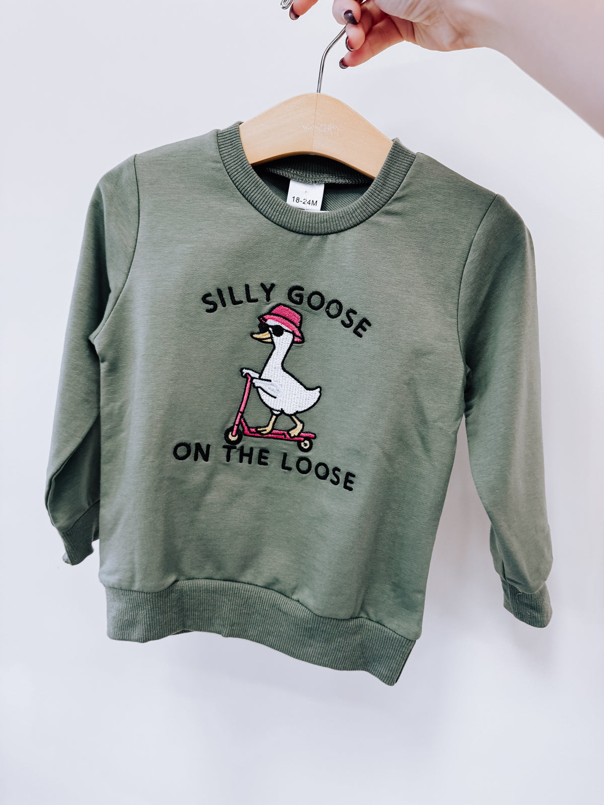 Tiny Tot Bundles Goose on the Loose Pullover, an olive green long-sleeve shirt featuring an embroidered goose in a pink hat riding a pink scooter with the text SILLY GOOSE ON THE LOOSE.