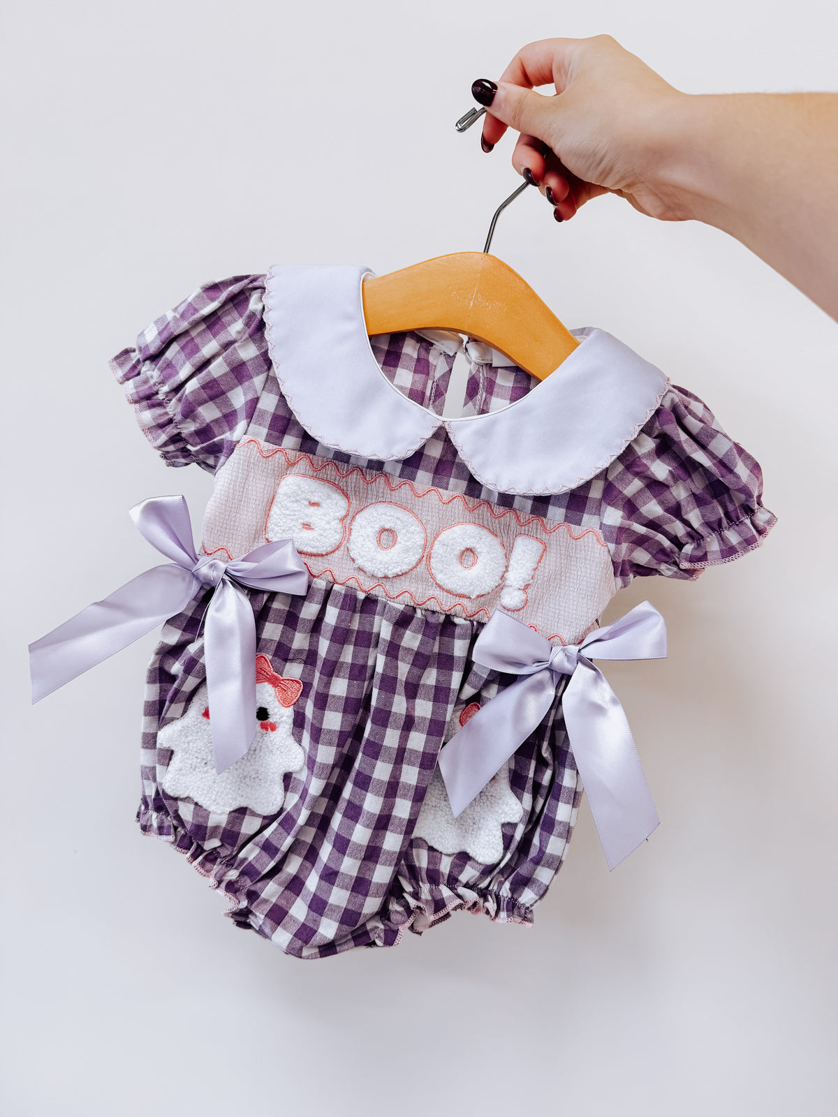 A hand holds a Tiny Tot Bundles Boo-tiful Purple Gingham Romper featuring a white collar, a BOO! embroidered chest panel, and two ghost appliqués.