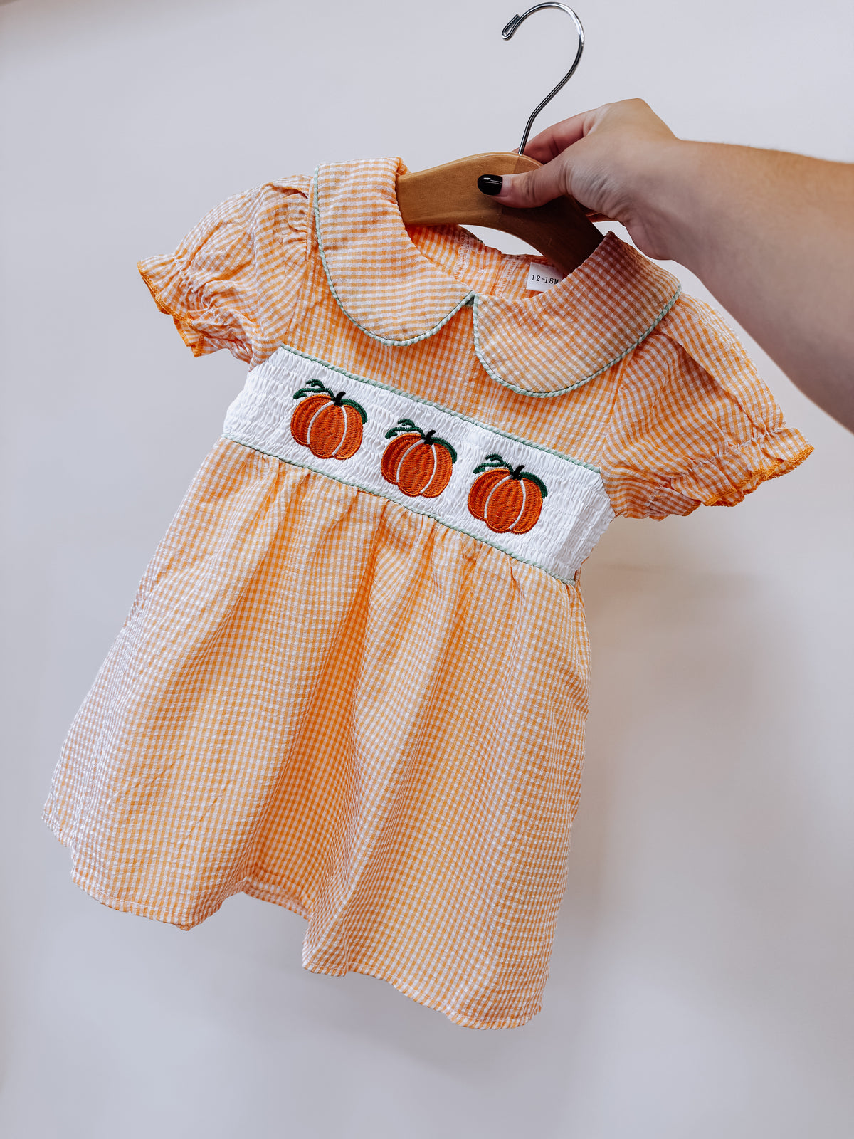 A hand holds a Tiny Tot Bundles Pumpkin Patch Smocked Dress, an orange and white gingham toddler dress with a smocked chest panel featuring three embroidered pumpkins.