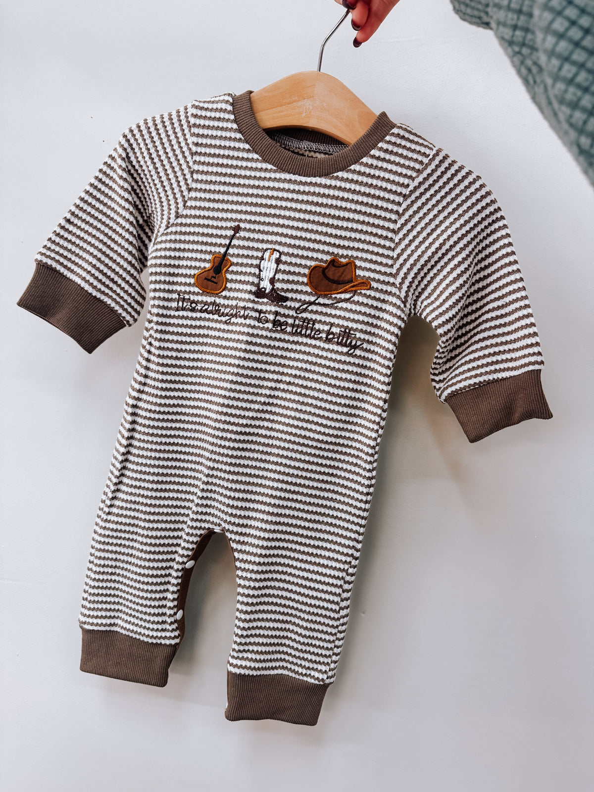Little Bitty Long Sleeve Romper with brown and white horizontal stripes, featuring embroidered icons of a guitar, a cowboy boot, and a cowboy hat above the text its alright to be little bitty.