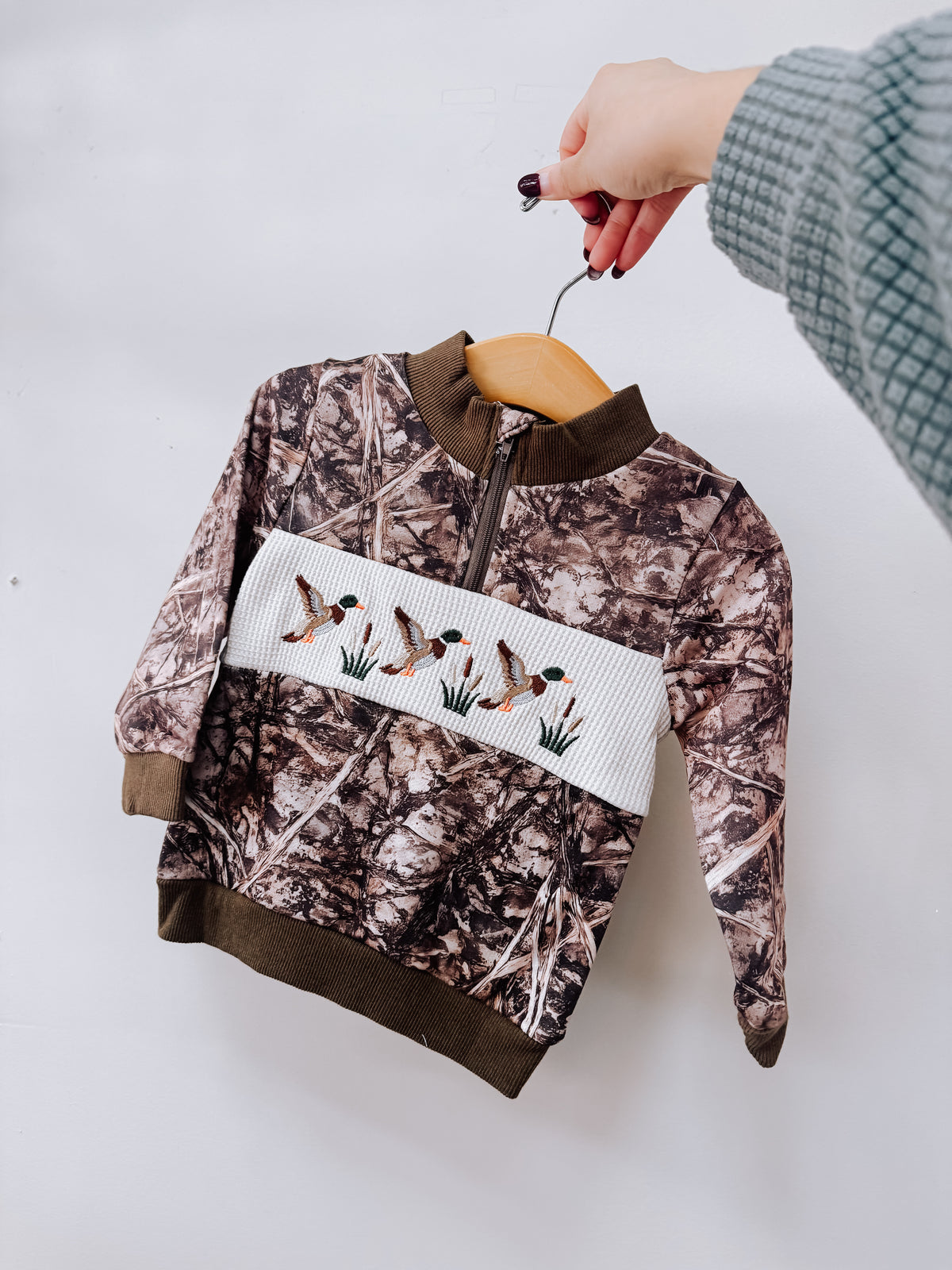 A Tiny Tot Bundles Camo Smocked Duck Pullover featuring a camo-print with a white smocked chest panel and embroidered mallard ducks, displayed on a wooden hanger.