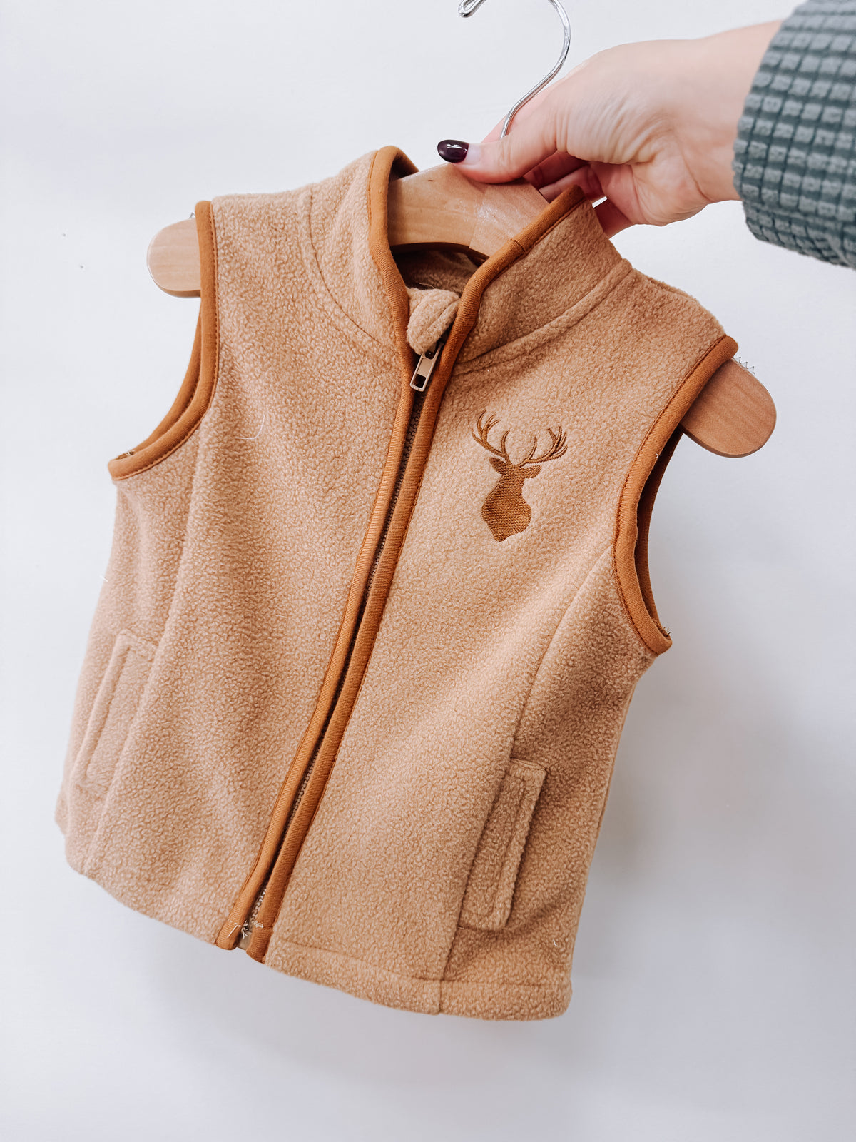 A Little Buck Vest in tan fleece with a brown zipper and an embroidered deer head on the chest, hanging on a wooden hanger.