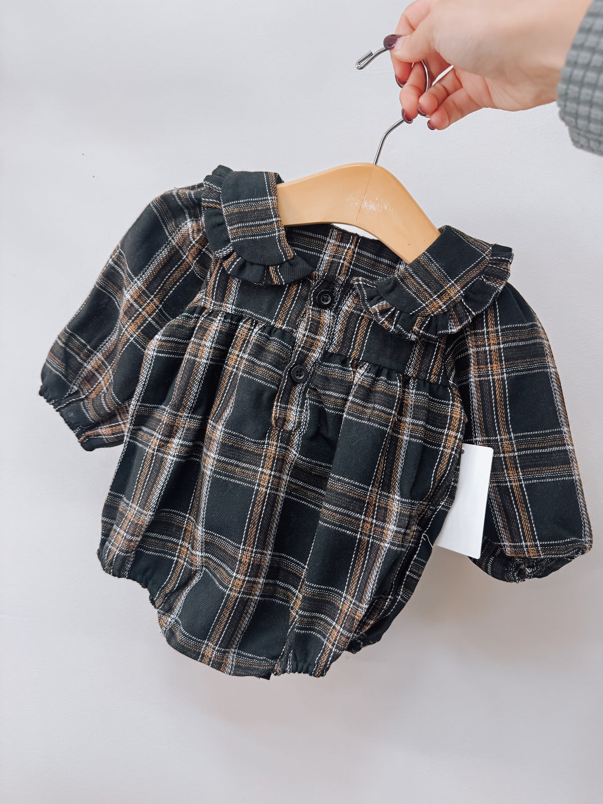 A Vintage Black Plaid Romper by Tiny Tot Bundles featuring a black and brown plaid pattern, puffed sleeves, and a ruffled collar, hanging on a wooden hanger.