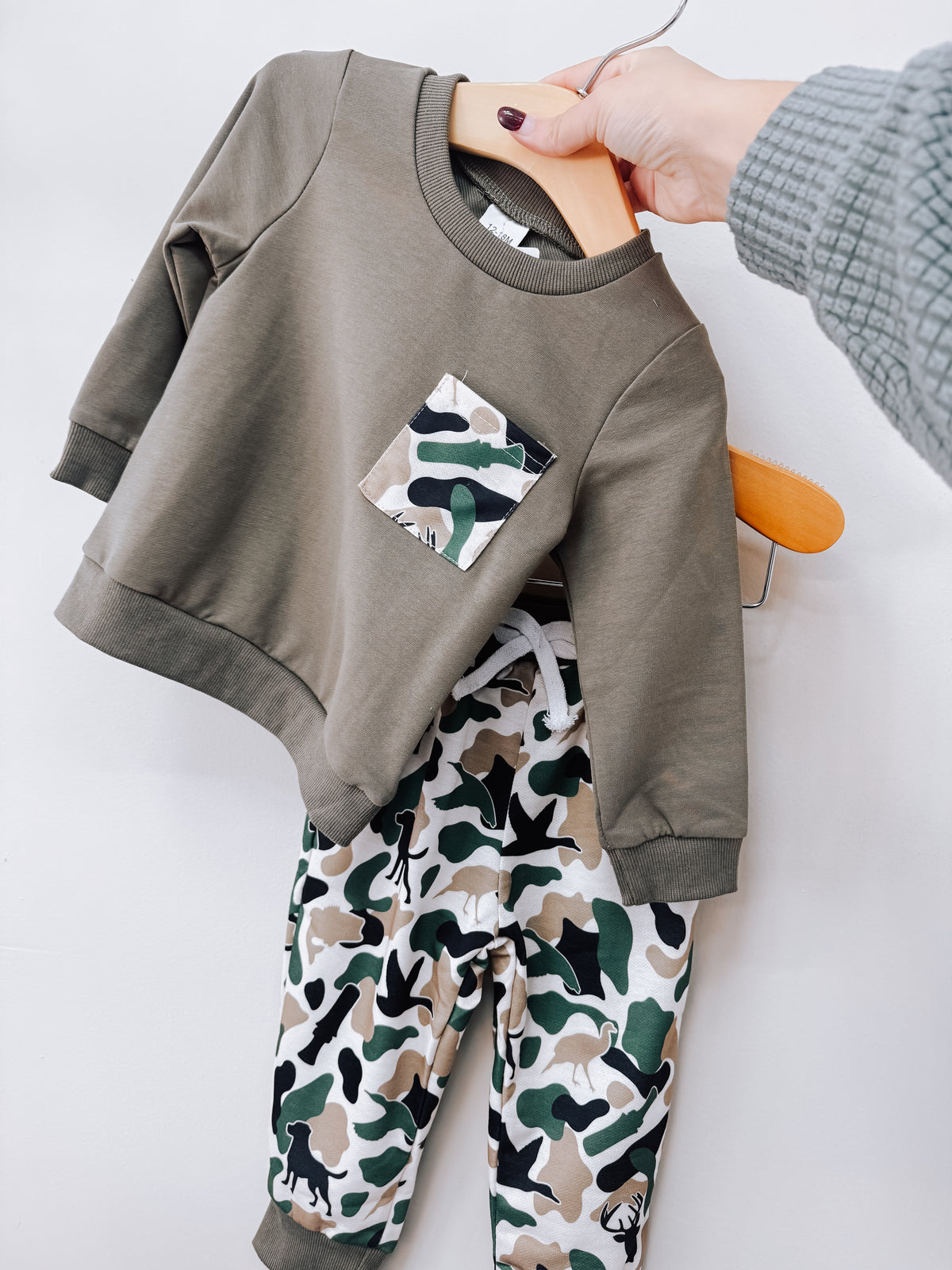 Tiny Tot Bundles Olive Camo Two-Piece Set consisting of a long-sleeved olive green pullover with a camouflage pocket and matching camouflage print joggers.