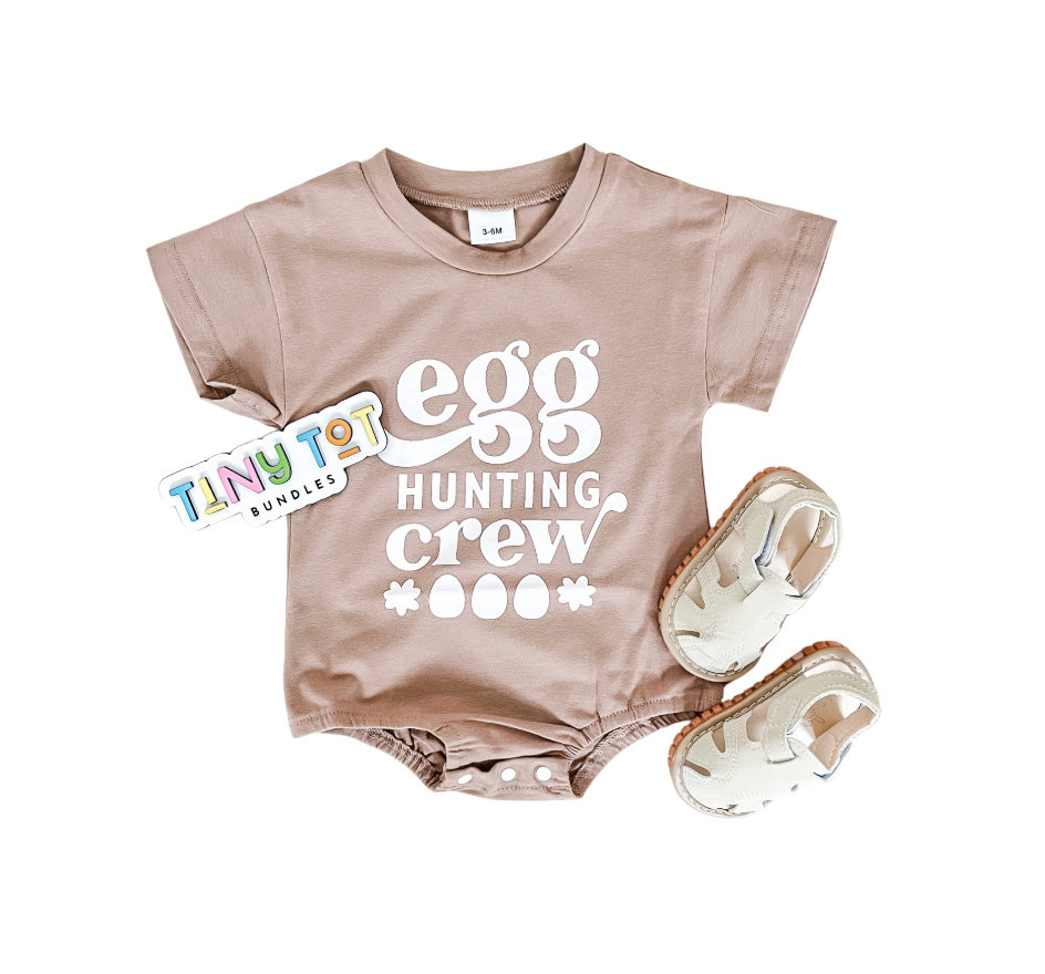 The Egg Hunting Crew Bubble by Tiny Tot Bundles is a light brown baby onesie with "egg hunting crew" in white, shown with beige baby sandals and a colorful branded tag on a white background.
