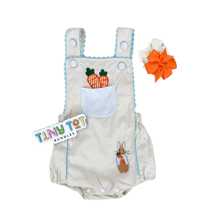 The Neutral Carrot Overall Romper by Tiny Tot Bundles is a beige romper with blue trim, a front pocket featuring two carrot appliqués and a bunny design near the leg, paired with white and bright orange hair bows.