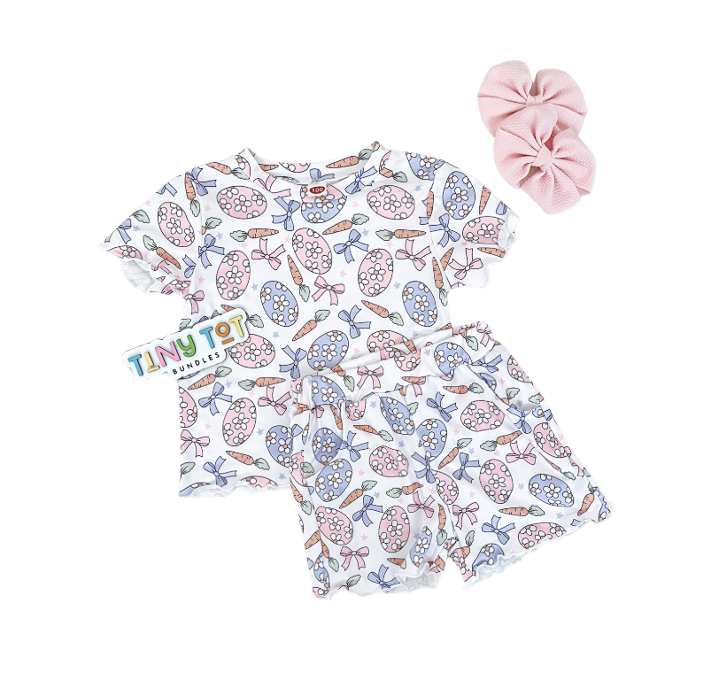 The Floral Egg Shorts Set by Tiny Tot Bundles features pastel Easter egg and bow prints, a light pink scrunchie, and a branded tag, all displayed on a white background.