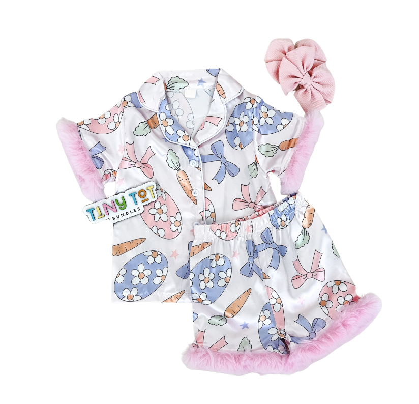The Floral Egg Pajama by Tiny Tot Bundles features a pastel print of bows, carrots, and flowers, pink faux fur trim, a matching pink scrunchie, and a branded tag attached to the pajama top.