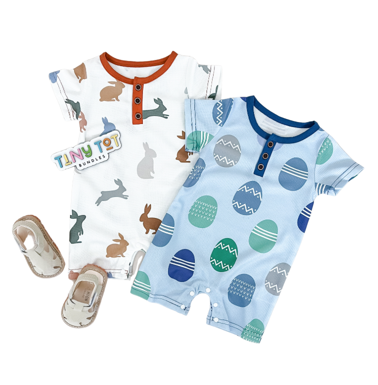 Two Infant Easter Jumpsuits by Tiny Tot Bundles are shown flat: one white with brown trim and bunny prints, the other blue with blue trim and Easter eggs. Tan baby sandals and a Tiny Tot Bundles label are also pictured.