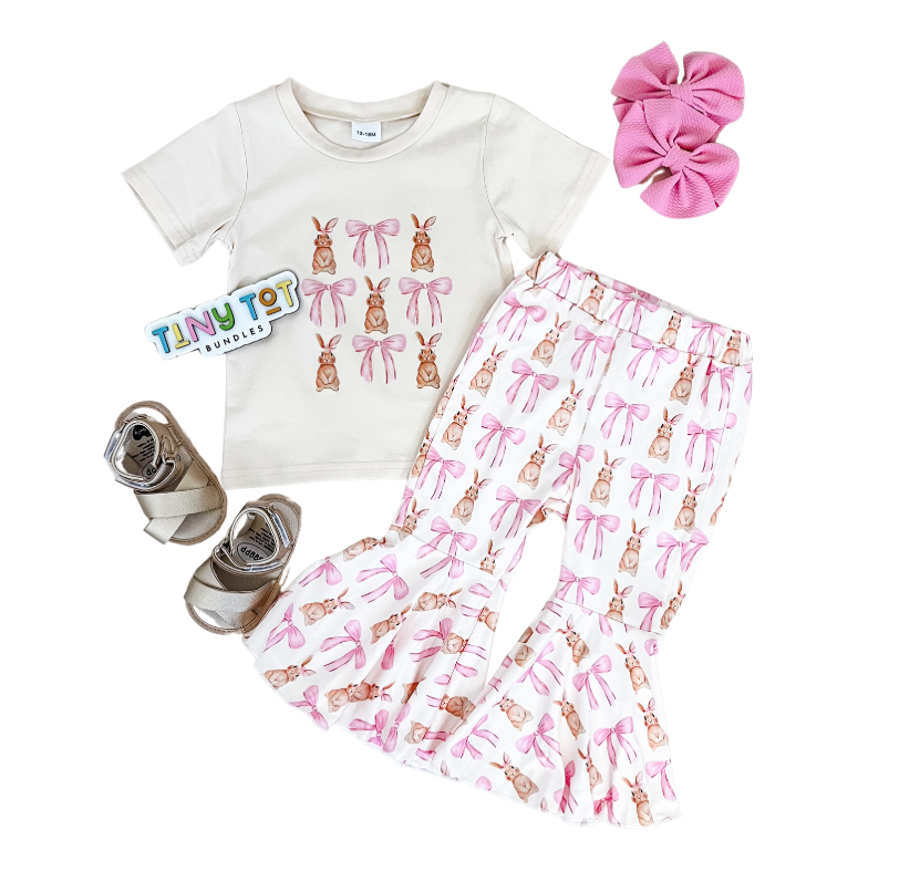 Flat lay of the Tiny Tot Bundles Coquette Bunny Bells Set, featuring a bunny print T-shirt and pink and white flared pants with bows, styled with metallic sandals, a pink hair bow, and a Tiny Tot Bundles sticker.