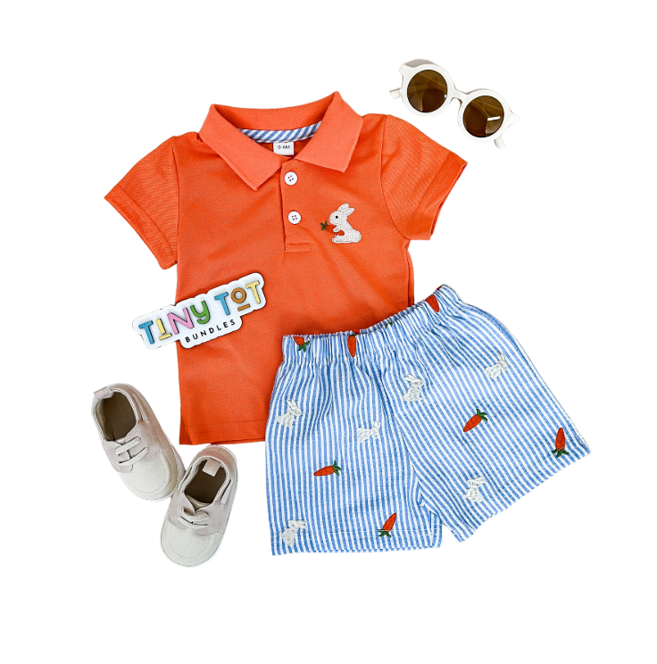 Flat lay of the Tiny Tot Bundles Easter Polo Shorts Set, showing an orange polo with a white bear patch, blue and white striped shorts with bunny and carrot designs, beige shoes, white sunglasses, and a brand sticker.