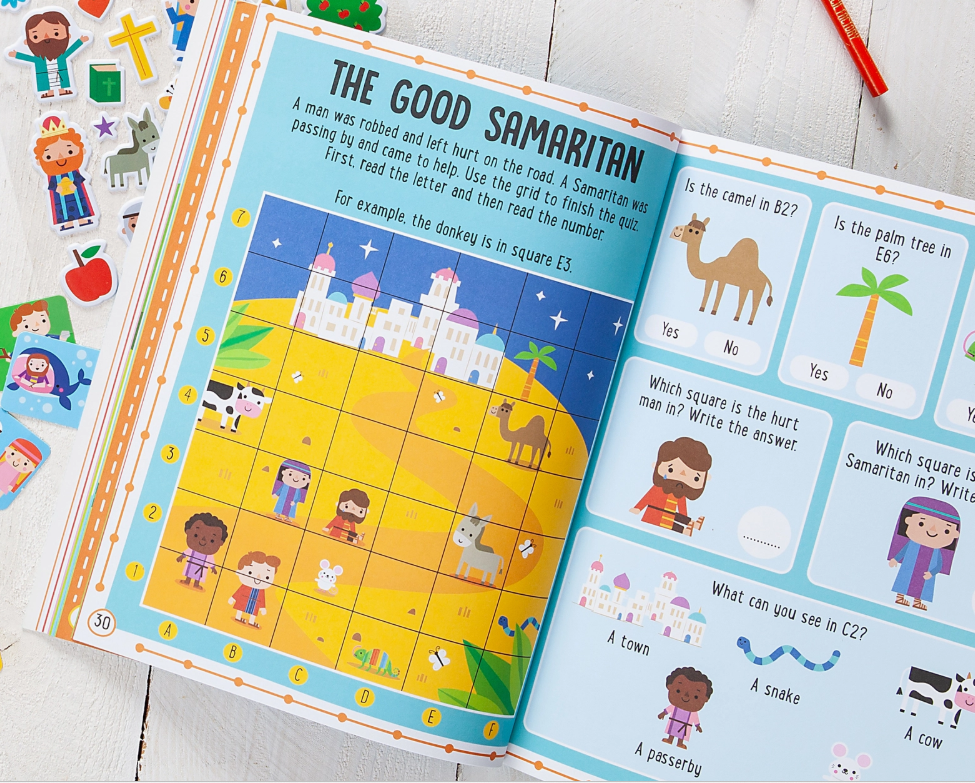 The My Bible Stories Activity Pink Book by Broad Street Publishing Group lies open, displaying a colorful Good Samaritan grid map puzzle with cartoon people, animals, and buildings. Stickers and a red pencil sit nearby on a white wooden surface.