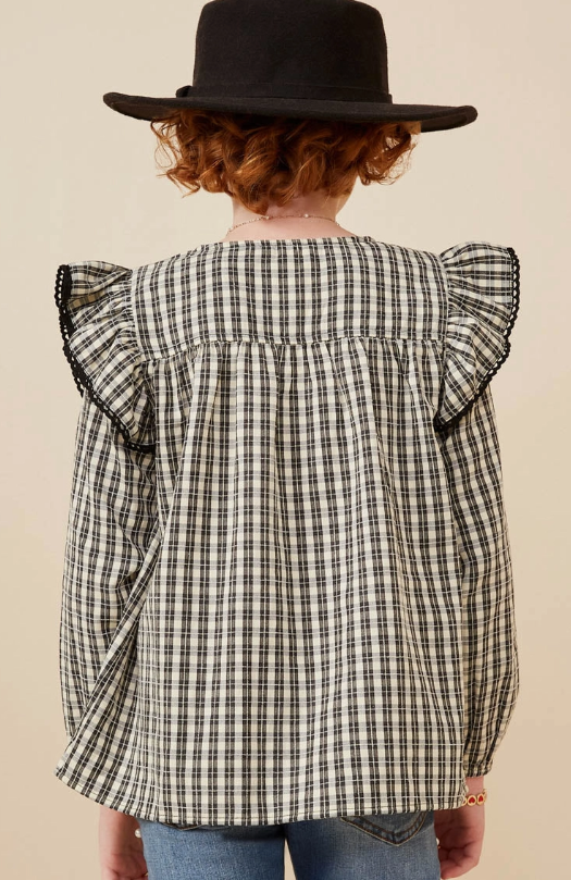 A child with curly red hair and a black hat stands facing away, wearing the Hayden LA Checkered Lace Trim Ruffled Shoulder Top and blue jeans.