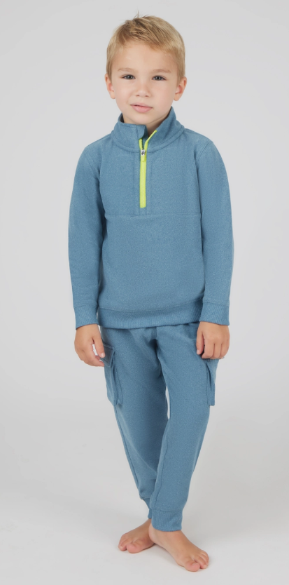 A young child in the 90 Degree by Reflex & Yogalicious Cozy Zip-Up Cargo Set—light blue fleece pullover with neon yellow zipper and matching cargo pants—smiles slightly, standing barefoot against a plain light background.