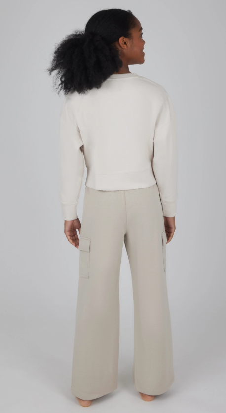 A person with curly hair in a ponytail stands barefoot, facing away, wearing the light long-sleeve top and wide-leg cargo pants from the 90 Degree by Reflex & Yogalicious Weekend Comfort Cargo Set.