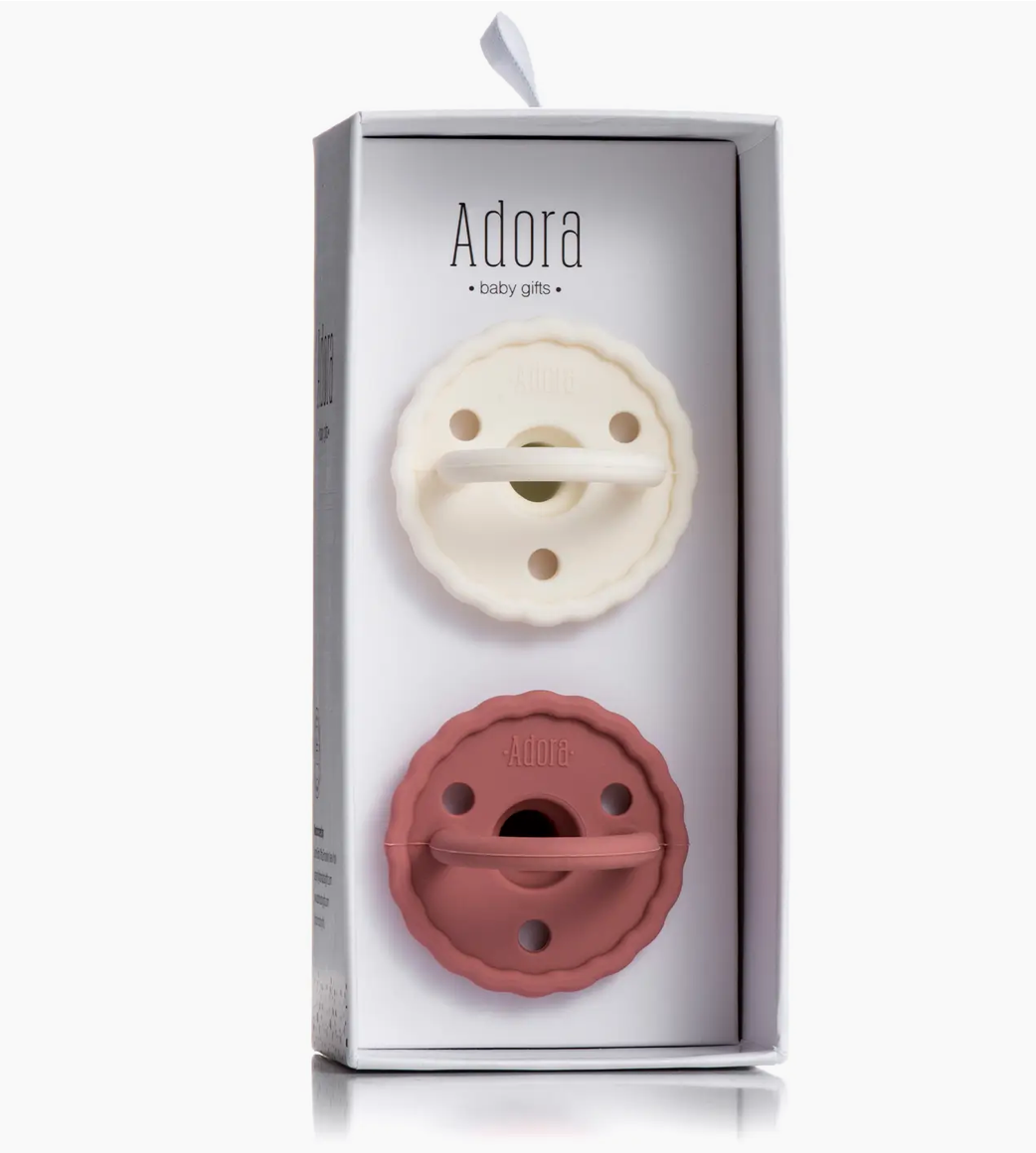 The Adora Saddle Adora Pacifier Set - Vanilla Rosewood features two pacifiers—one cream, one mauve—with a round, scalloped-edge design, packaged vertically in a white box.
