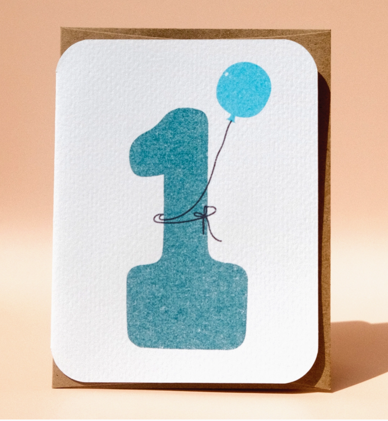 The 1st Birthday Card by Paper Chain Studios features a large blue number 1 with a small blue balloon on a light background.