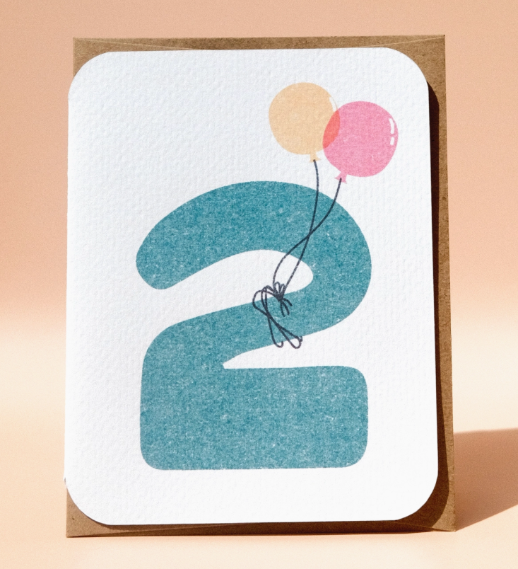 The 2nd Birthday Card by Paper Chain Studios features a large blue number 2, peach and pink balloons attached by black string, rounded corners, and a brown backing.