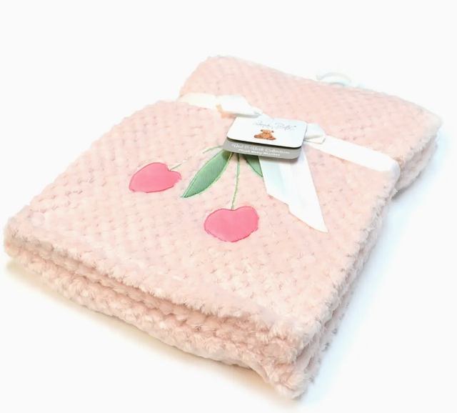 The Cherry Sweet Snuggle Blanket by Lollipop Dreams is a soft, light pink textured baby blanket with two embroidered pink cherries, neatly folded and tied with a white ribbon. A product tag is attached.
