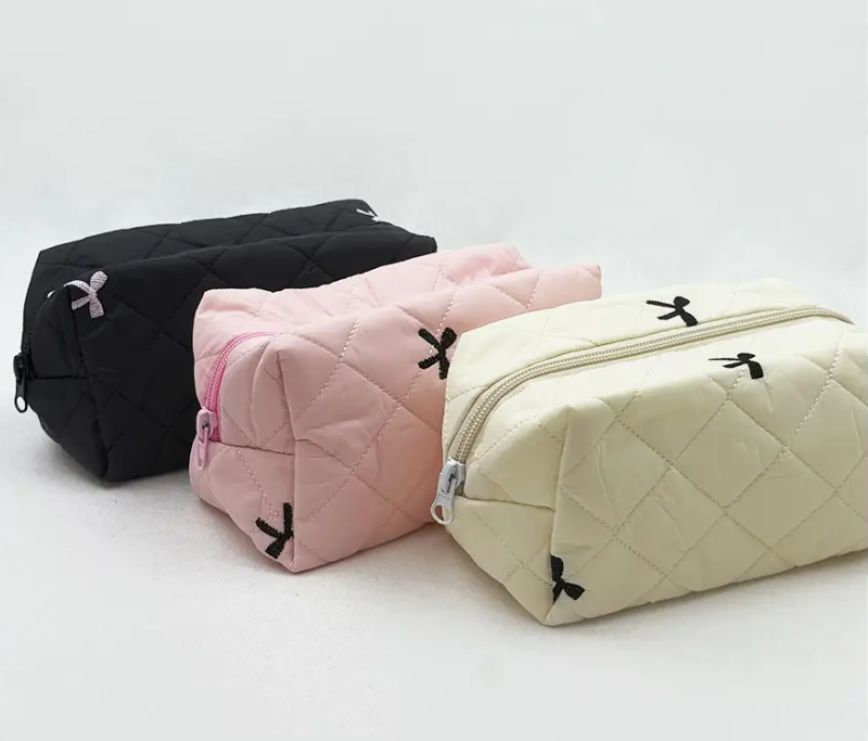 Three love & repeat Petite Bow Quilted Pouches in black, pink, and cream—each accented with a small corner ribbon—are displayed on a simple white background.