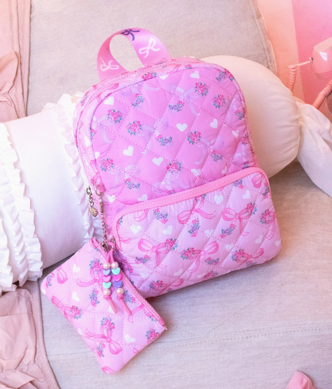 The omg Accessories Sweetheart Bows Quilted Backpack, featuring heart and bow designs, comes with a matching pouch and pastel beaded keychain, shown on a light couch with ruffled white pillows in a softly lit room.