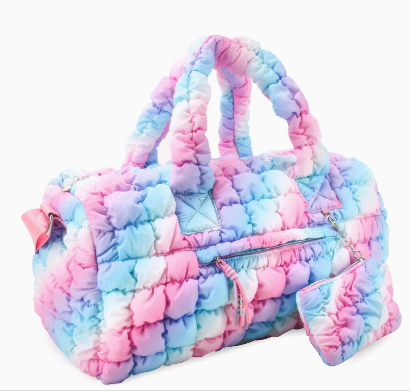 The Pastel Puff Dream Bag by omg Accessories is a quilted duffel with a pink, blue, and purple pastel tie-dye pattern, padded handles, a front zipper pocket, and an attached matching pouch.