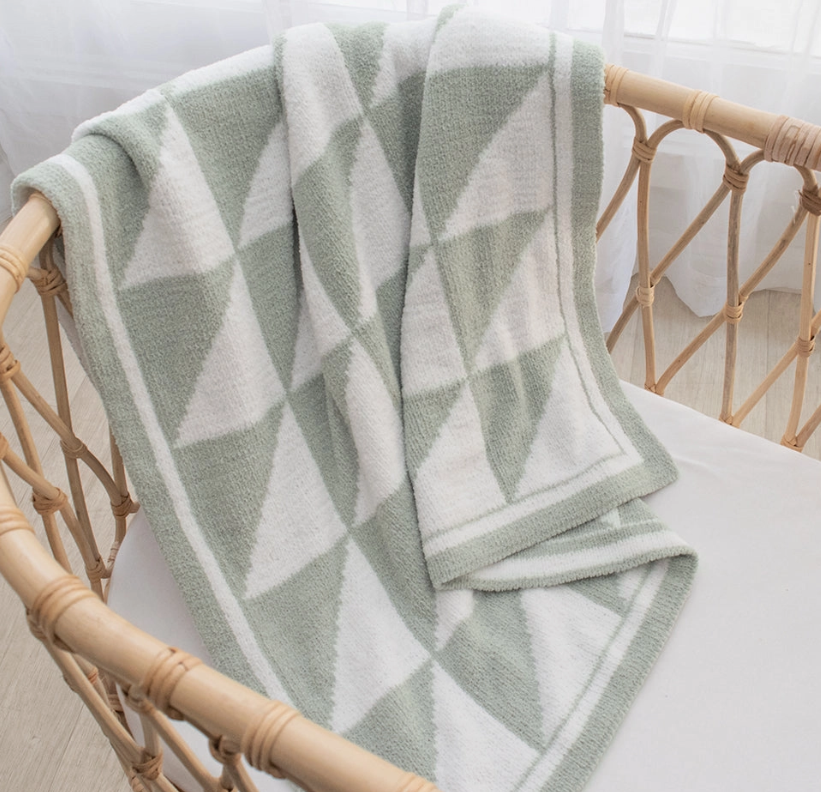 The living textiles Chenille Baby Blanket - Sage Triangle, featuring a green and white geometric pattern, is draped over a light wooden crib with rattan sides as natural light filters through a sheer curtain in the background.