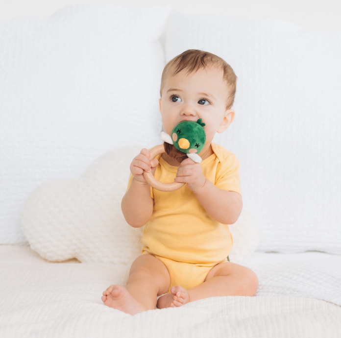 A baby in a yellow onesie sits on a white bed, holding and chewing on the PearHead Snuggle Duck Baby Teether, while looking to the side.