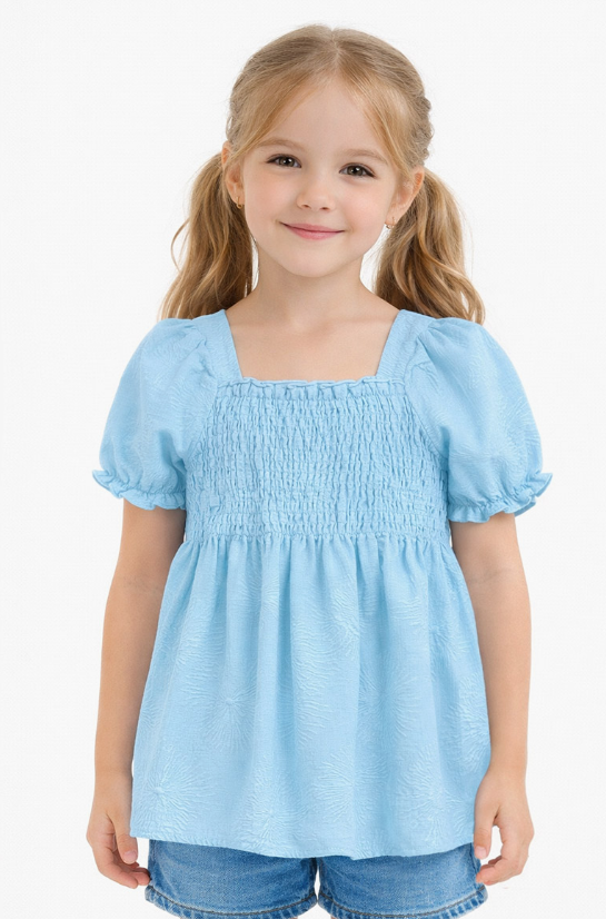 A girl with light brown pigtails smiles in the Cutie Patootie Sky Blossom Smocked Top and denim shorts, standing against a plain white background.