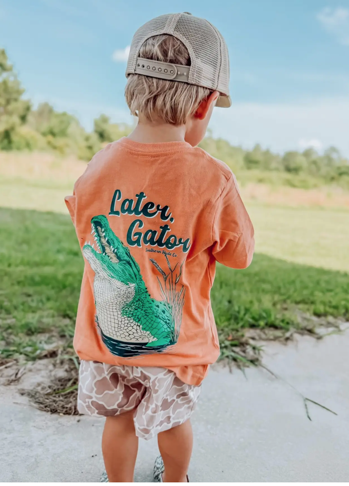 A young child in a Wild Trail Outfitters Later Gator Toddler Tee and beige cap stands outside on a sunny day, facing away from the camera toward a grassy field.