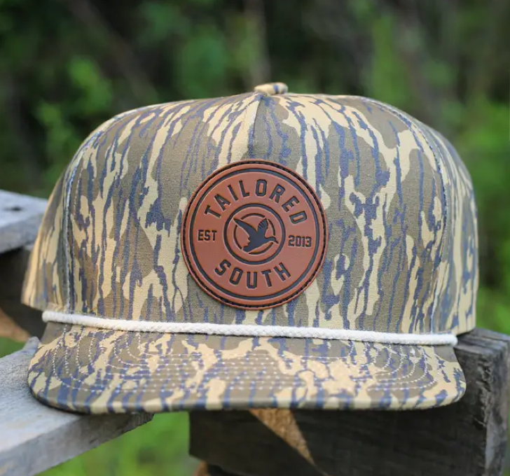 The Lowland Tan Rope Duck Leather Patch Adult Hat by Wild Trail Outfitters features a camouflage pattern, round brown leather patch with TAILORED SOUTH EST 2013 and bird logo, plus a white rope detail above the brim.