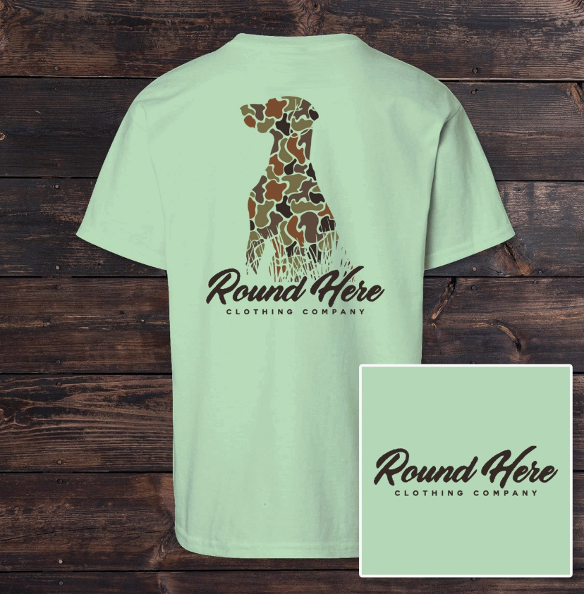 The Old North State's Hunting Dog Youth Tee features a light green color with a camo-patterned dog silhouette and brand text on the back, plus matching text on the front left chest. Shirt shown on a wooden background.