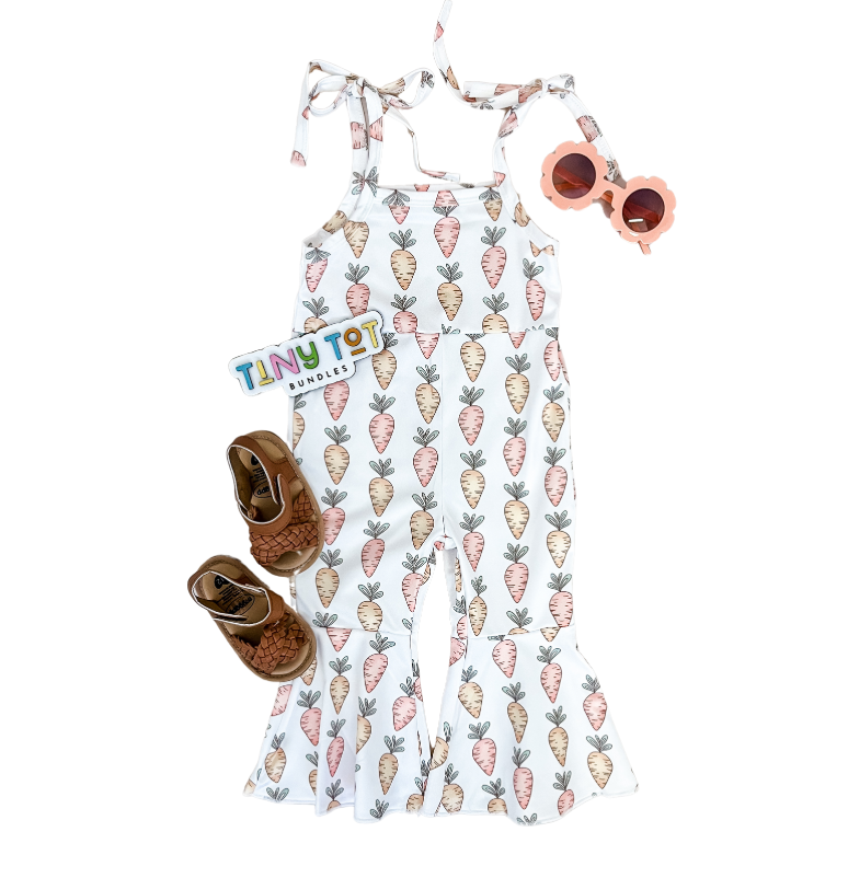 Flat lay of a toddler’s outfit: Tiny Tot Bundles Carrot Easter Jumpsuit, brown woven sandals, pink flower sunglasses, and a Tiny Tot Bundles sticker arranged neatly on a white background.