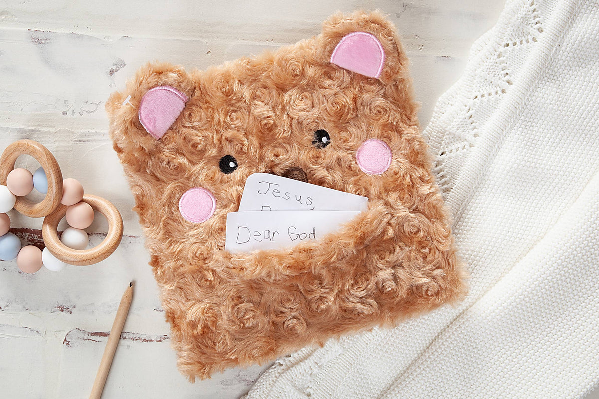 A BroadStreet Publishing Group, LLC Prayer Bear Book rests on a light wooden surface, accompanied by a pencil, teething rings, and a soft white textured blanket.