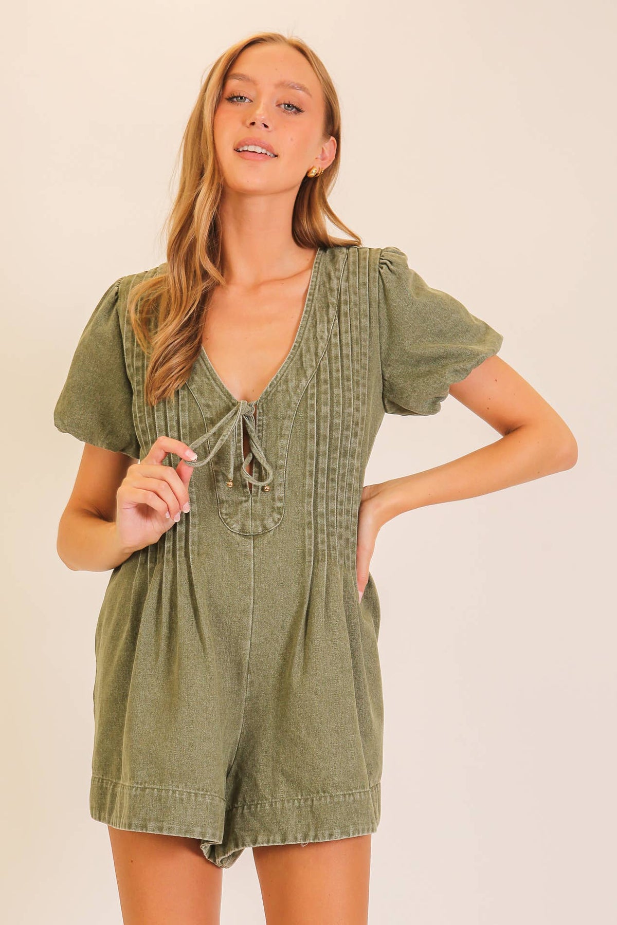 A woman poses against a plain background wearing the Lumiere Desert Muse Utility Romper in olive green, featuring short sleeves, a tie neckline, vertical stitching details, and her right hand on her hip.