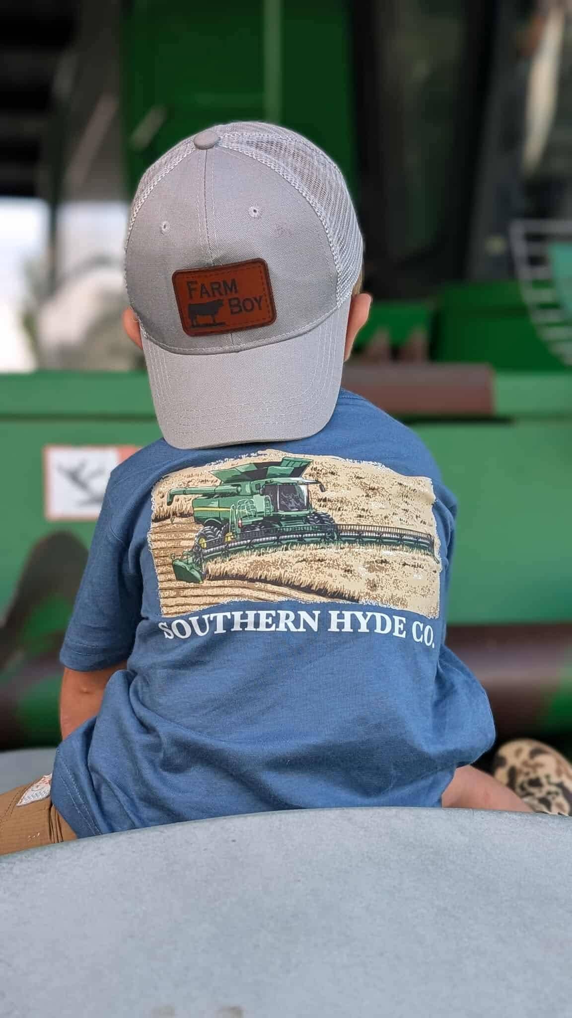 A young child seen from behind wears a gray baseball cap and the Southern Hyde Co. Grain Train Tee, a blue t-shirt with a graphic of a combine harvester in a field.