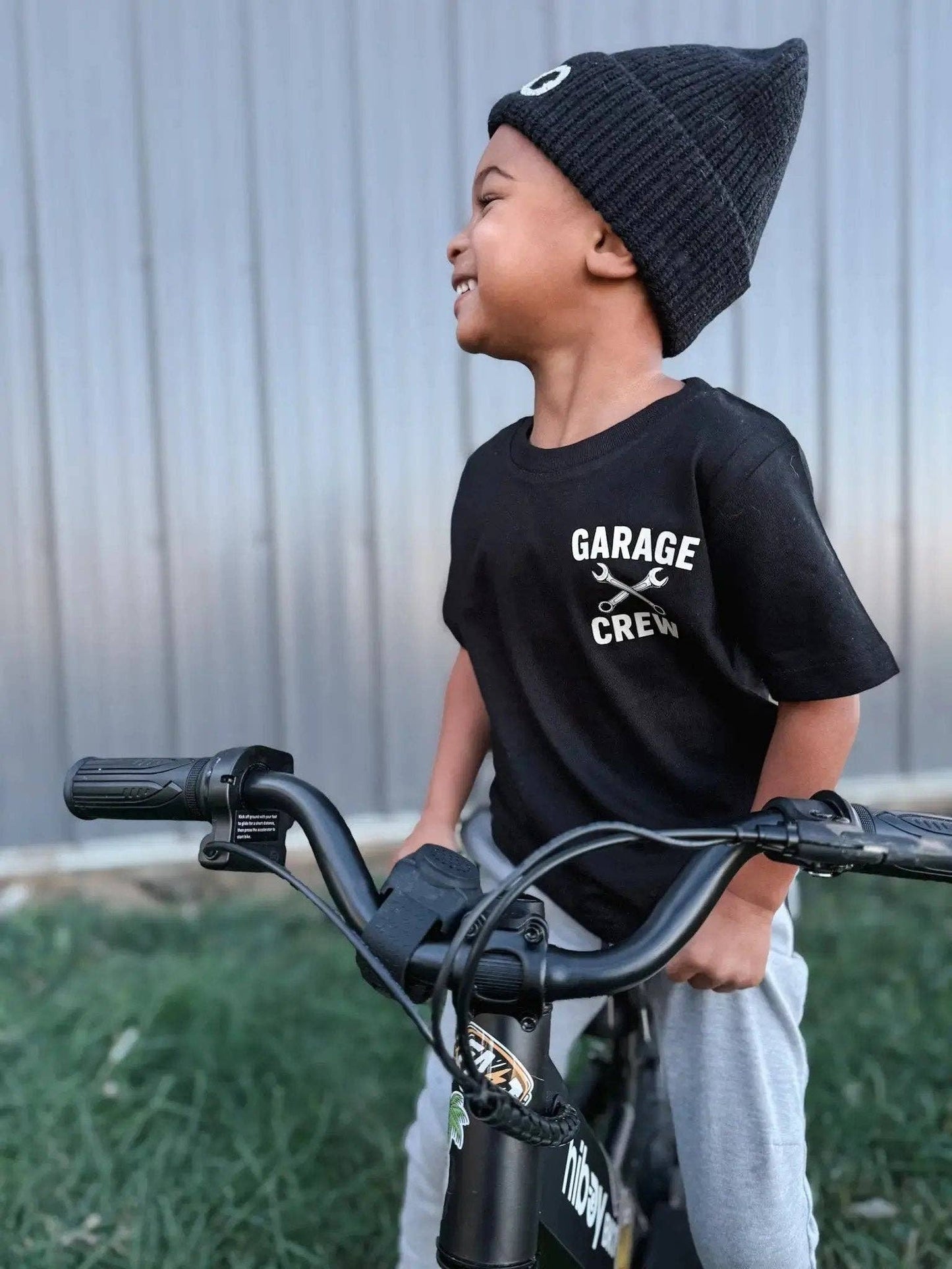 A smiling child wearing a black beanie and the Raising Brave "My Dad's Garage" Toddler Tee sits on a bike outdoors, looking to the side with a corrugated wall in the background.