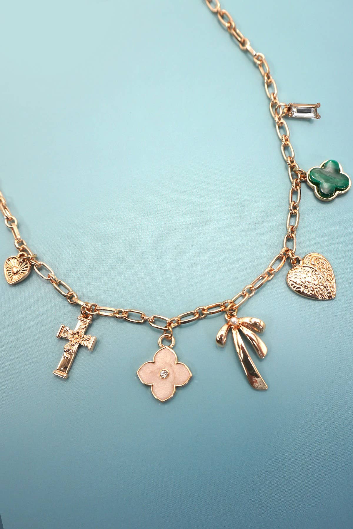 The Gold Charm Necklace from JOSSLYN by wall to wall, featuring a heart, cross, flower, bow, and green clover charm, is displayed on a light blue background.