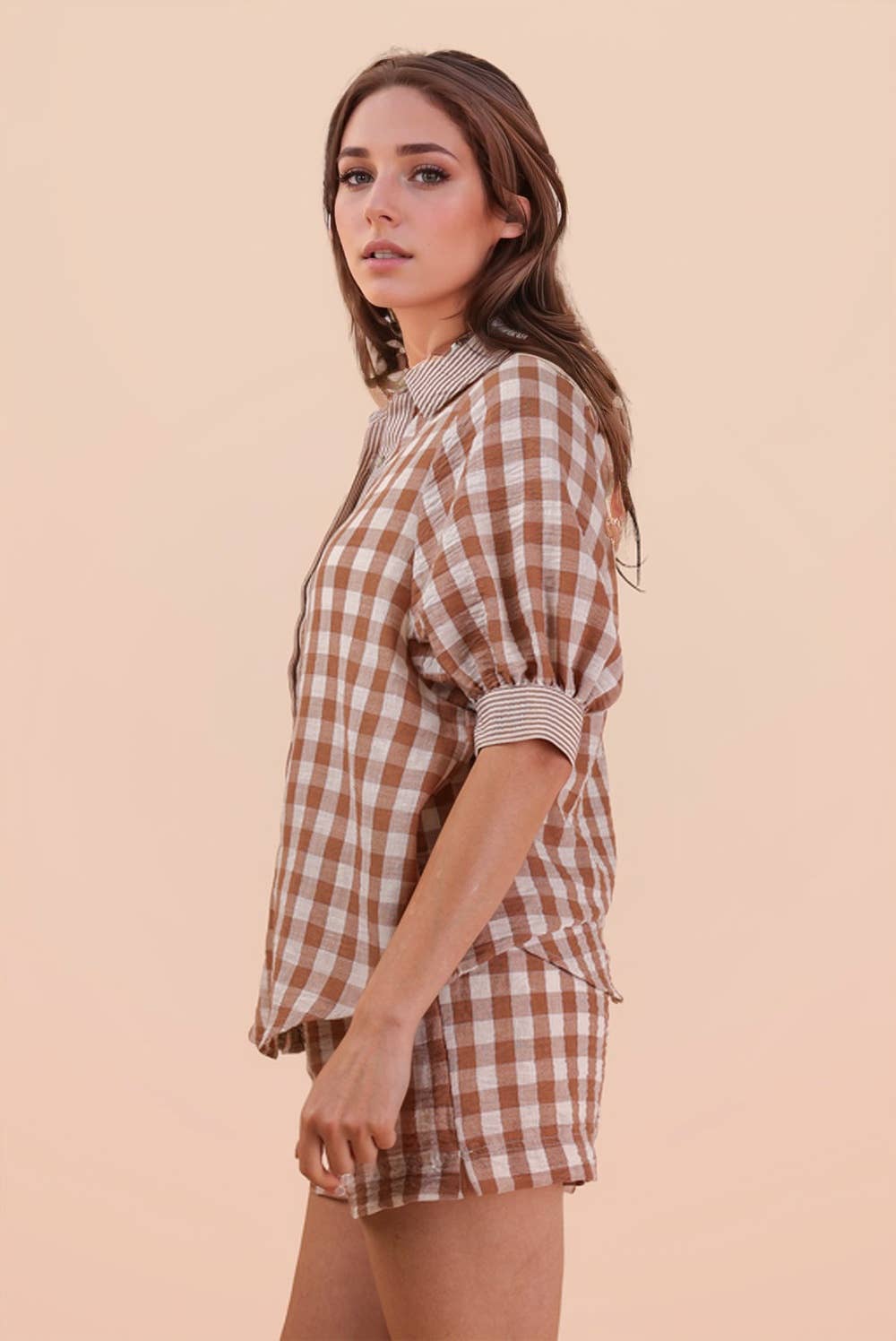 A woman stands sideways against a plain beige background, wearing the VERY J Checkered Shirt Top & Shorts Set featuring a brown and white plaid pattern with rolled sleeves.