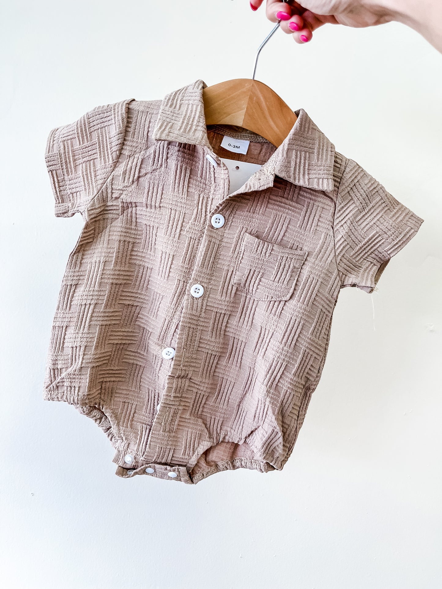 A hand holds a hanger displaying the Tiny Tot Bundles Sandstone Textured Bubble— a beige, short-sleeve baby romper with a checkered texture, white buttons, collar, and small chest pocket against a plain white background.