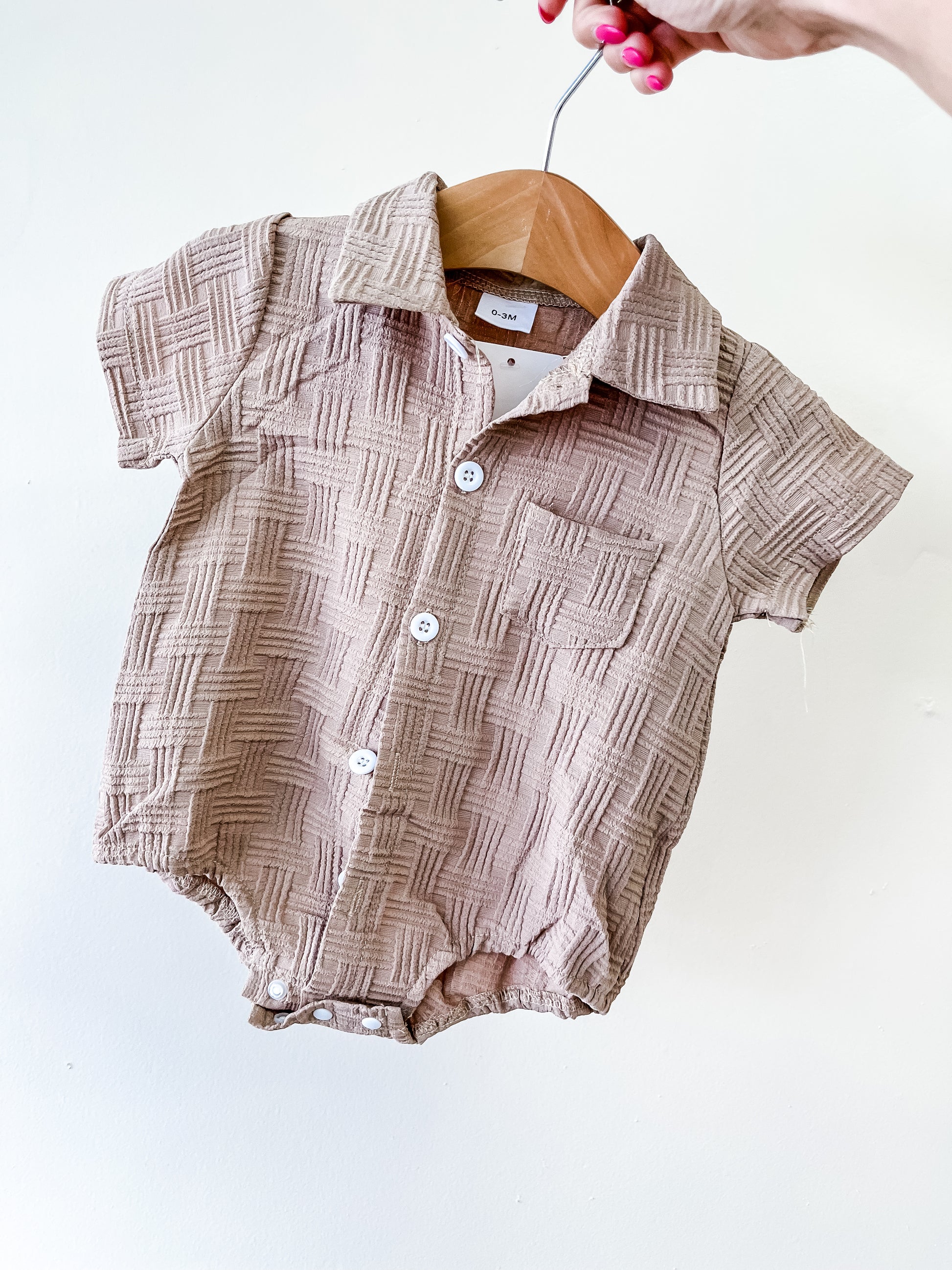 A hand holds a hanger displaying the Tiny Tot Bundles Sandstone Textured Bubble— a beige, short-sleeve baby romper with a checkered texture, white buttons, collar, and small chest pocket against a plain white background.