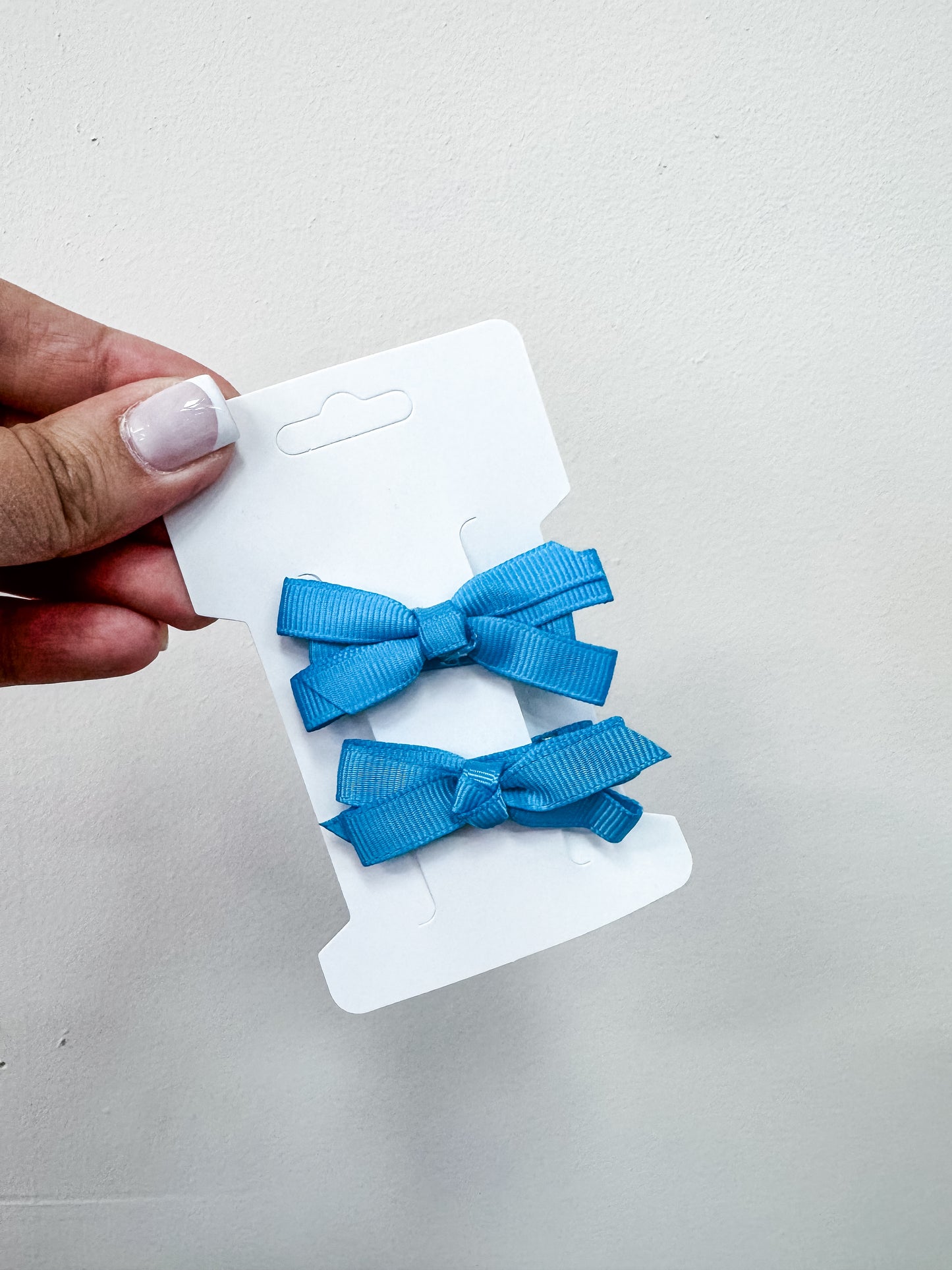 A hand holds a white card displaying the Tiny Tot Bundles Duo Piggy Clip Pack—two blue ribbon hair bows—against a plain, light background.