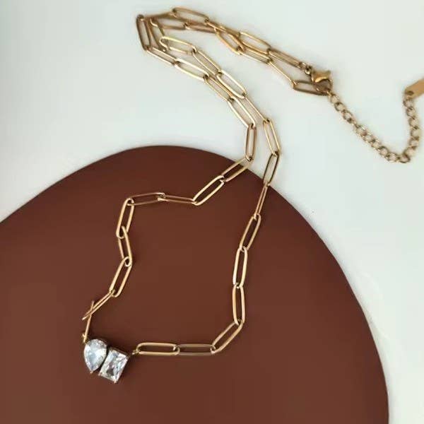 A Gold Rhin Necklace by JOSSLYN by wall to wall, featuring a gold paperclip chain and two clear gemstones, rests on a white and brown surface.