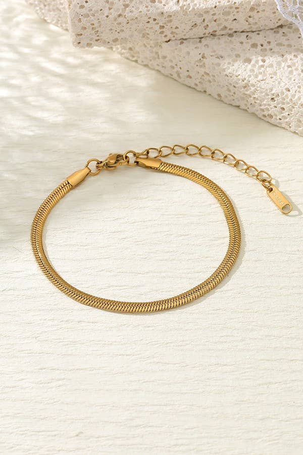 The Dainty Gold Bracelet by JOSSLYN from wall to wall features a sleek, adjustable snake chain with an extender, elegantly displayed on a softly textured surface with delicate lace fabric partially visible in the background.