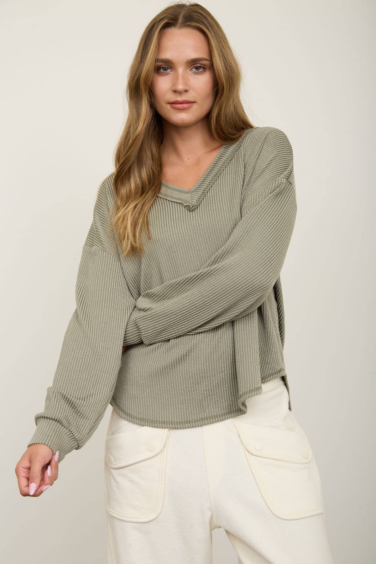 A woman with long, wavy hair wears the Blu Pepper Olive V-Neck Long Sleeve Top paired with cream-colored, high-waisted pants featuring large pockets, standing against a plain light background.