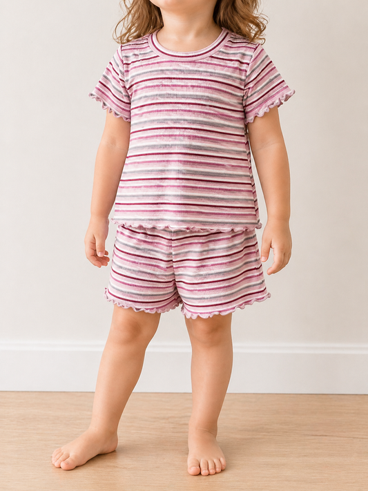 A young child stands barefoot indoors wearing the Tiny Tot Bundles Rosy Stripe Snuggle Set, a matching short-sleeve top and shorts with pink, purple, and white stripes plus ruffled edges. The child’s head is not fully visible.