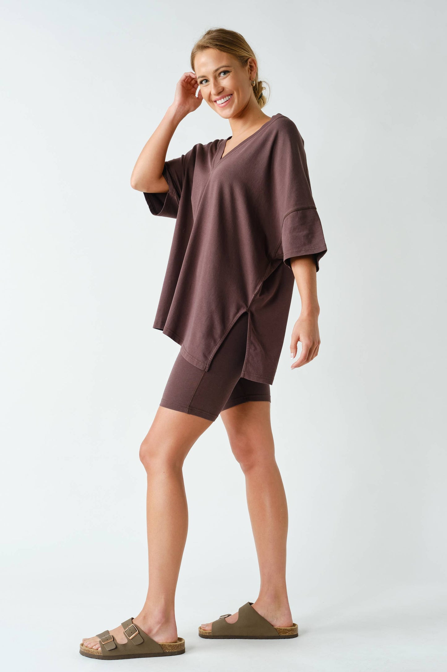 A woman stands smiling against a plain white background, wearing the Mono B Switchback Pima Cotton Set—a loose brown oversized top paired with matching biker shorts—and tan sandals.